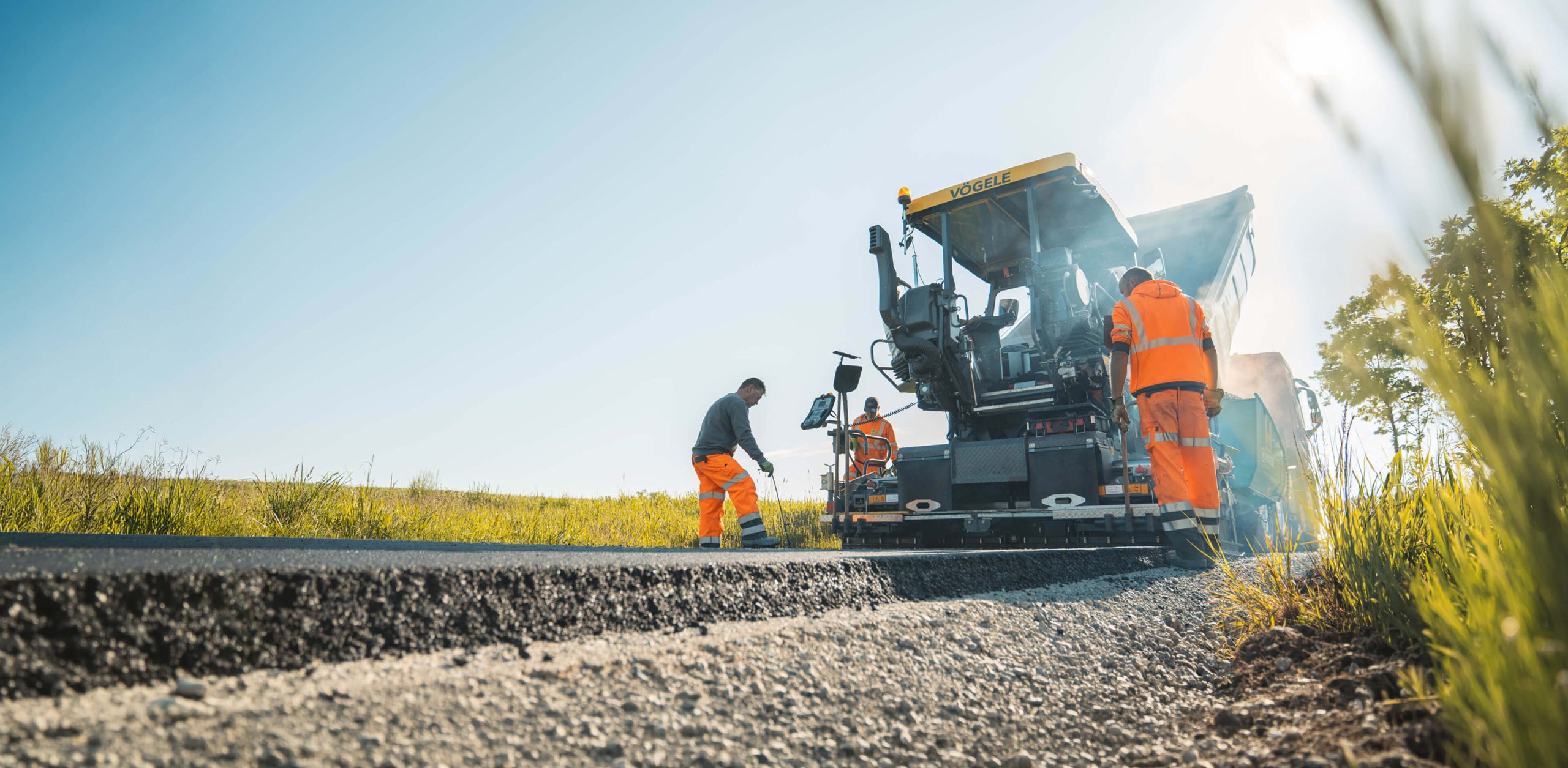 Mitarbeiter von Christian Knoch Tiefbau beim Einbau von Asphalt mit einem Straßenfertiger auf einer ländlichen Straße.