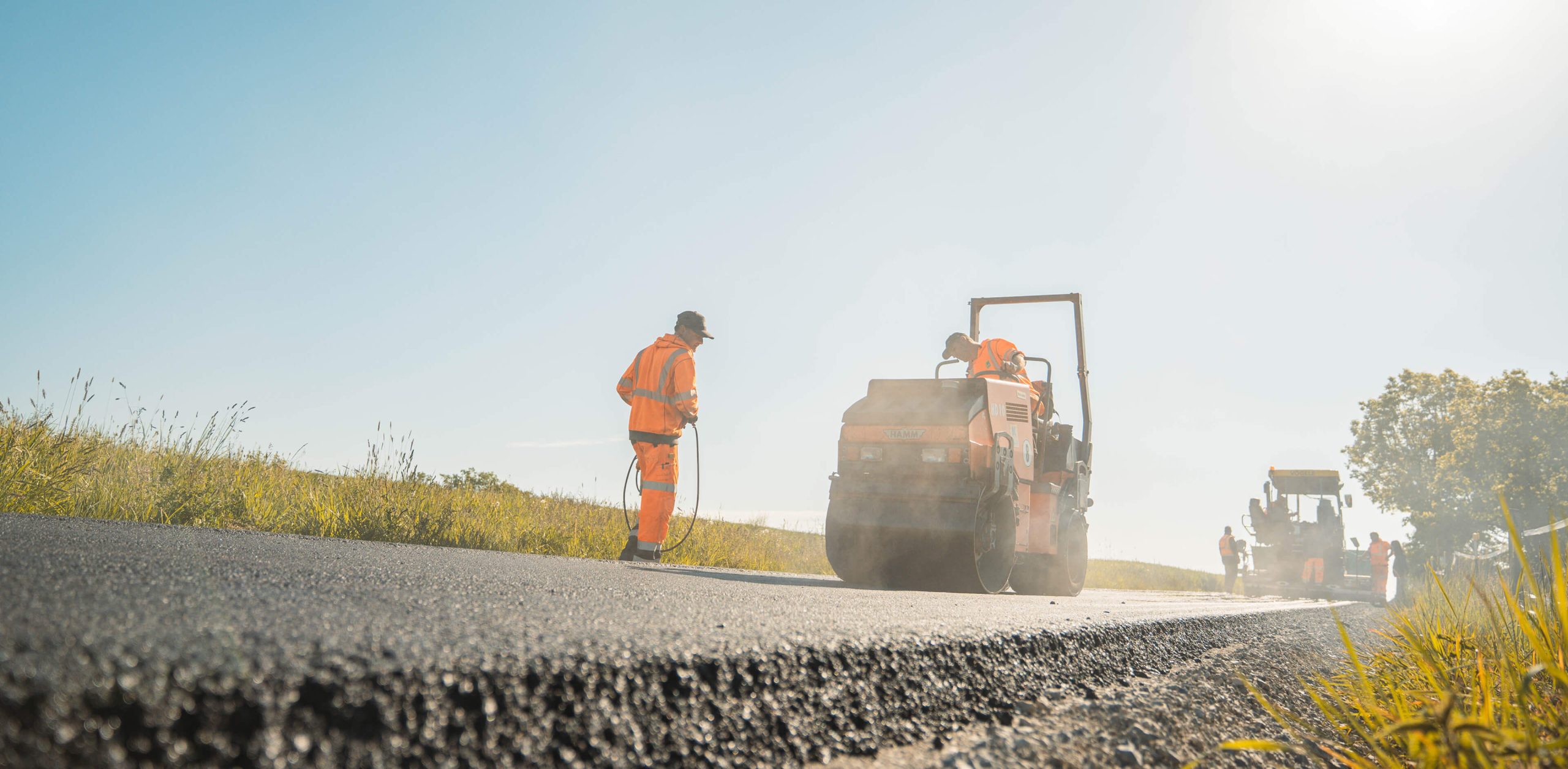 Straßenbaustelle unter blauem Himmel: Mitarbeiter von Christian Knoch Tiefbau verdichten frisch eingebauten Asphalt mit einer Straßenwalze.