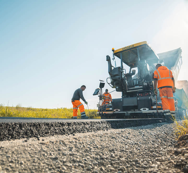 Mitarbeiter von Christian Knoch Tiefbau beim Einbau von Asphalt mit einem Straßenfertiger auf einer ländlichen Straße.