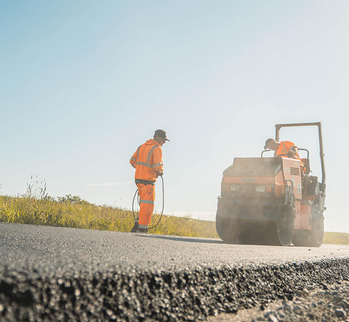 Straßenbaustelle unter blauem Himmel: Mitarbeiter von Christian Knoch Tiefbau verdichten frisch eingebauten Asphalt mit einer Straßenwalze.