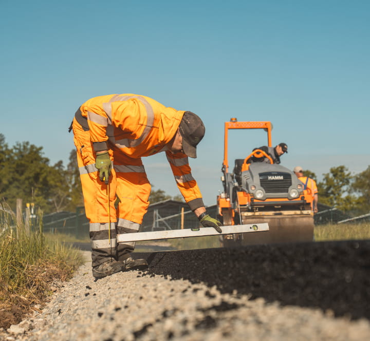 Aufnahme eines Mitarbeiters von Christian Knoch Tiefbau bei Kontrollarbeiten mit Wasserwaage vor einer Straßenwalze – Ausbildung im Straßenbau.